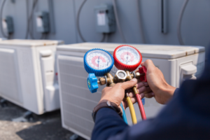 Squeaks technician holds a manifold gauge set with red and blue dials near outdoor air conditioning units.