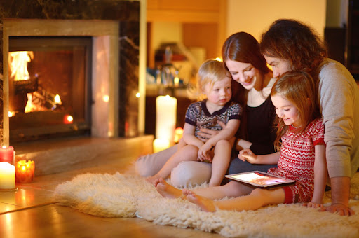 A family of four sitting on the floor near a fireplace. They're looking at a tablet.