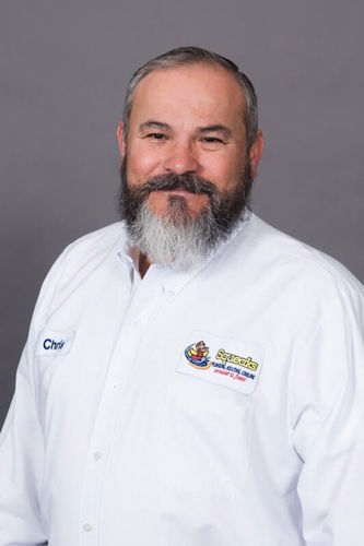 A middle-aged man with a gray beard wearing a white button-up shirt with embroidered Squeaks logo and a "Chris" name tag, standing against a plain gray background.