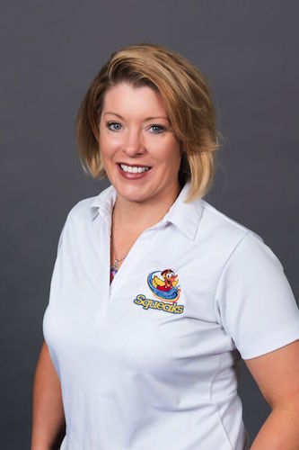 Woman with short blonde hair smiling, wearing a white polo shirt with a "Squeaks" cartoon logo, standing against a plain gray background.