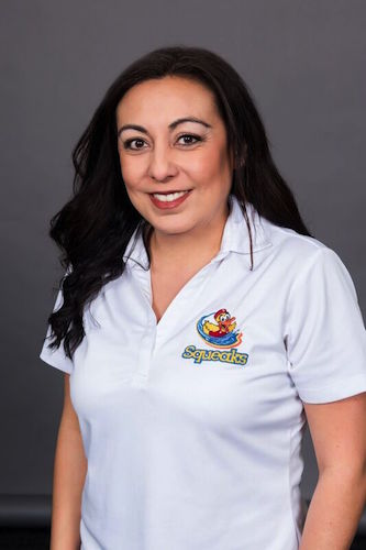 Woman with long dark hair wearing a white polo shirt with a colorful "Squeaks" logo, standing in front of a plain dark background, smiling at the camera.