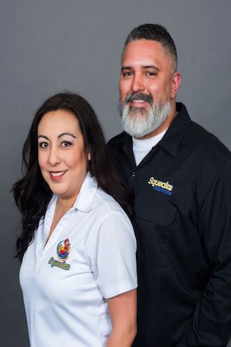 A woman and a man stand side by side, both wearing collared shirts with embroidered Squeaks logos, in front of a plain gray background.
