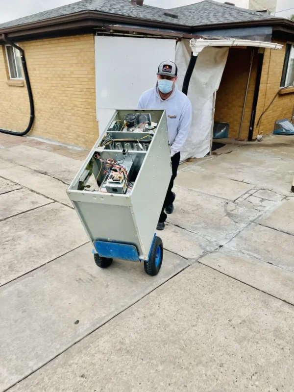 A person wearing a mask and cap moves a large, upright electronic device on a blue hand truck along a concrete driveway outside a building.