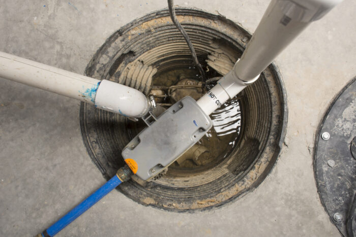 Open sump pit with water, containing a submersible sump pump and multiple connected pipes, set in a concrete floor.