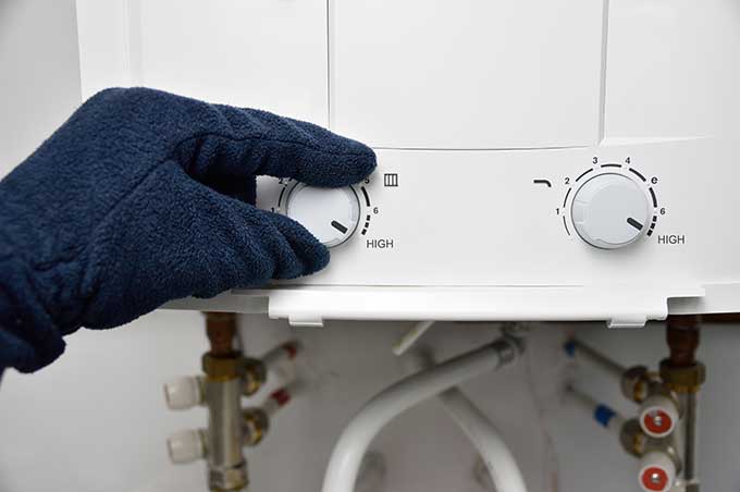 A gloved hand adjusts the temperature control knob on a water heater, with visible pipes and valves below the unit.