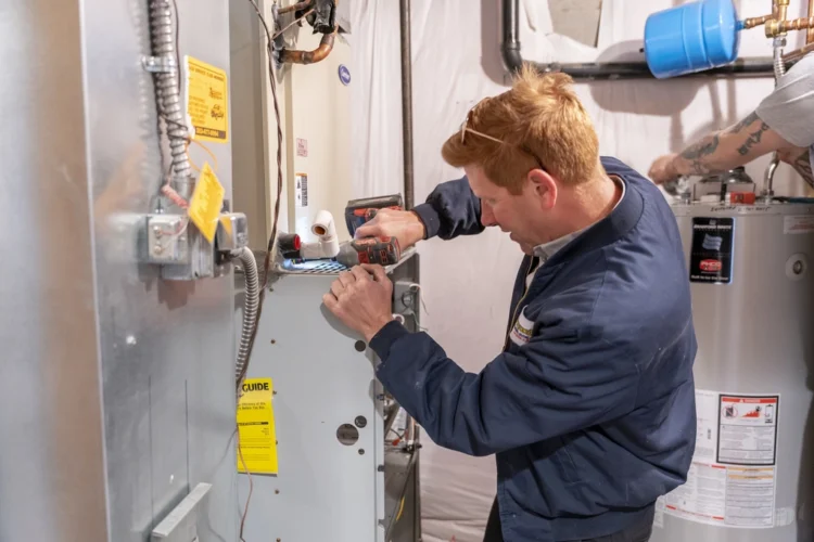 A technician in a navy jacket uses a power drill to service or repair a furnace in a basement utility room.