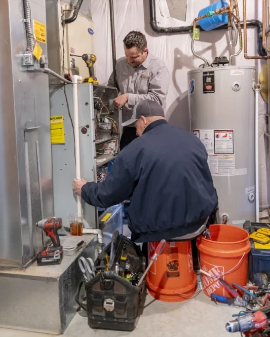 Two techs conducting furnace maintenance, with one technician seated on an orange bucket servicing the unit
