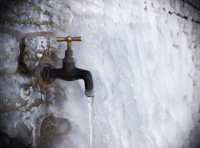 A metal outdoor faucet with a small stream of water is surrounded by thick ice on a wall, indicating freezing temperatures.