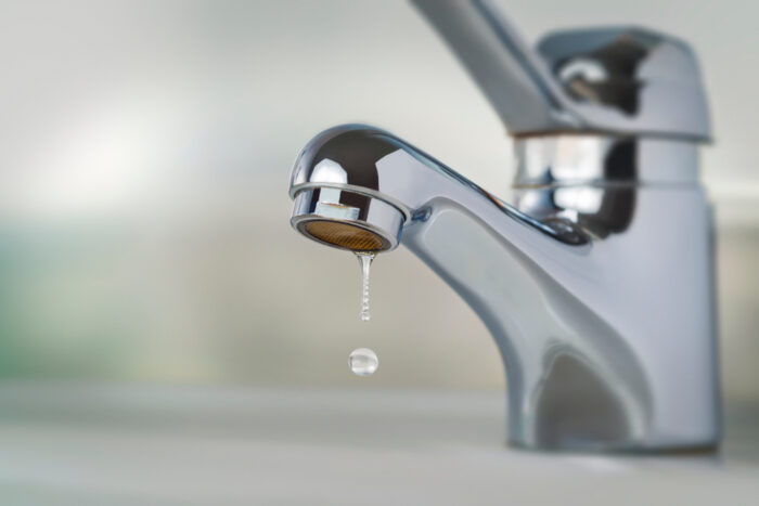A close-up of a chrome faucet with a single drop of water falling from the spout against a blurred background.