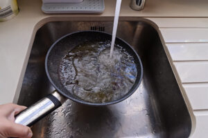 Hand holding pan with cooking grease under running water in sink.