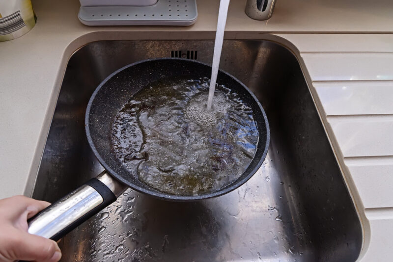 A persons hand holding a frying pan with cooking grease under running water in kitchen sink.