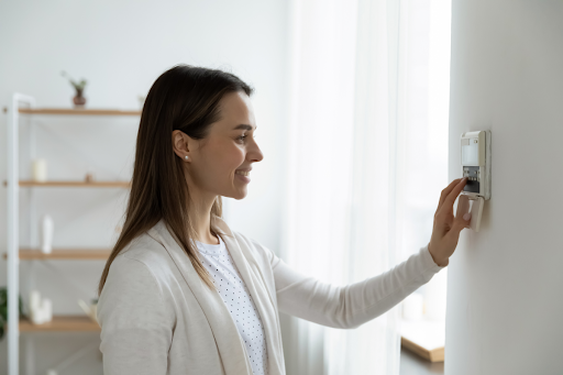 A woman stands indoors adjusting a wall-mounted thermostat. She is smiling and wearing a white cardigan, with shelves and a window visible in the background.