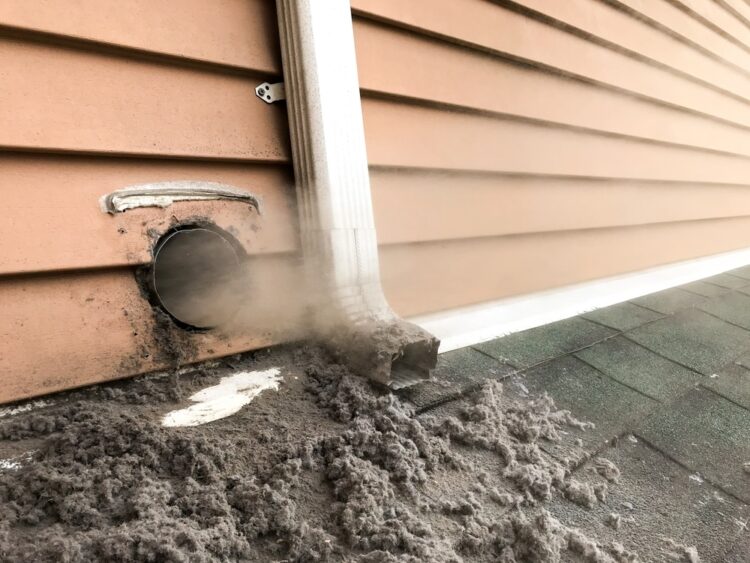 Dryer vent on the exterior wall of a house clogged with lint and debris, with dust spilling onto the roof shingles below.