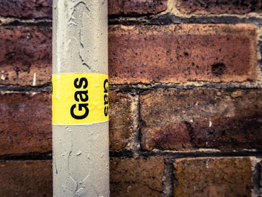 A metal pipe with a yellow label that reads "Gas" is installed in front of a brick wall.