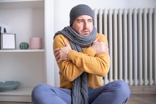 man with winter clothes on inside his home shivering in front of broken furnace